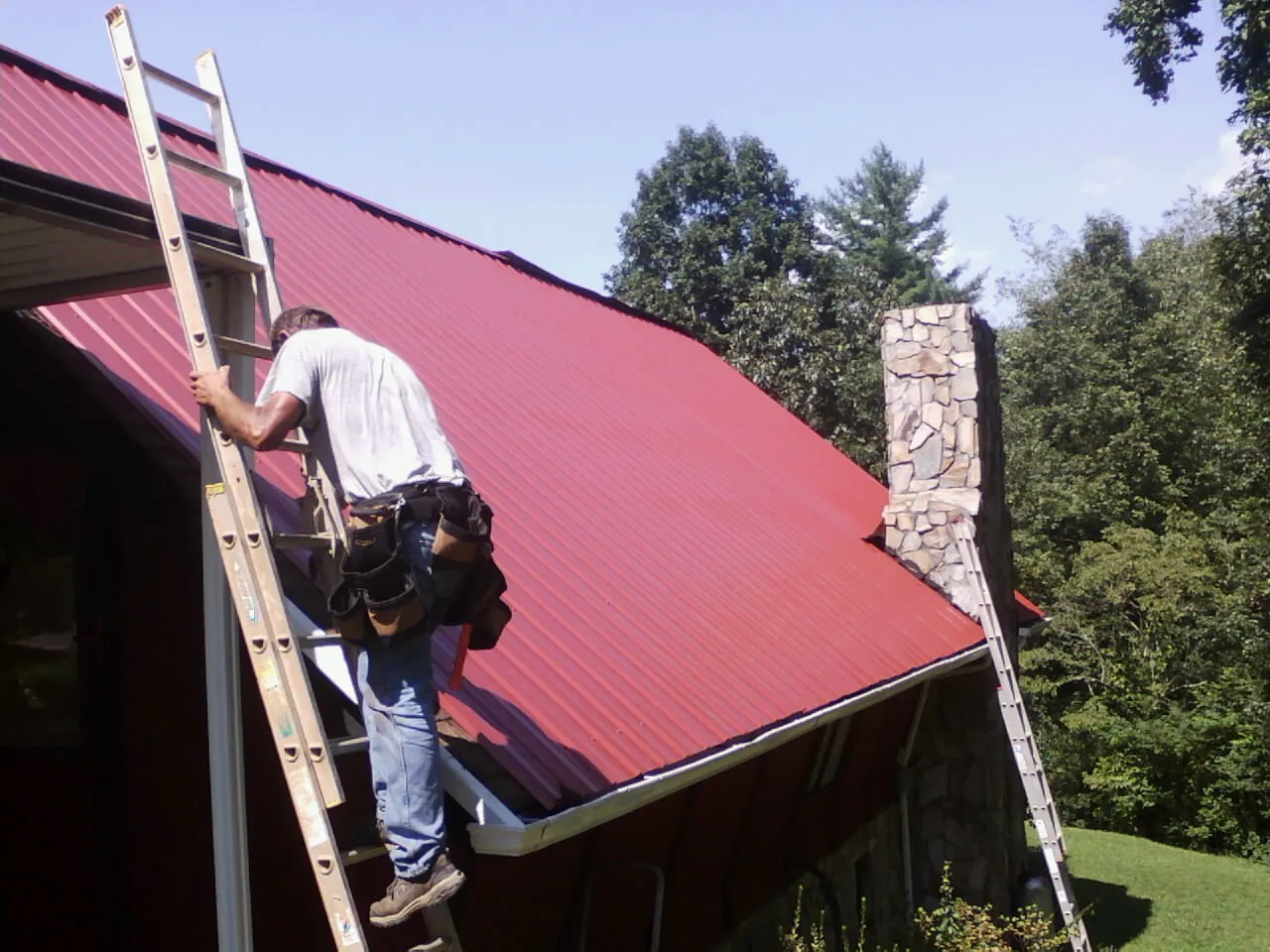Roofer on a ladder beside a red metal roof and stone chimney — Soffit & Fascia Repair by Roofing Brothers in Walton